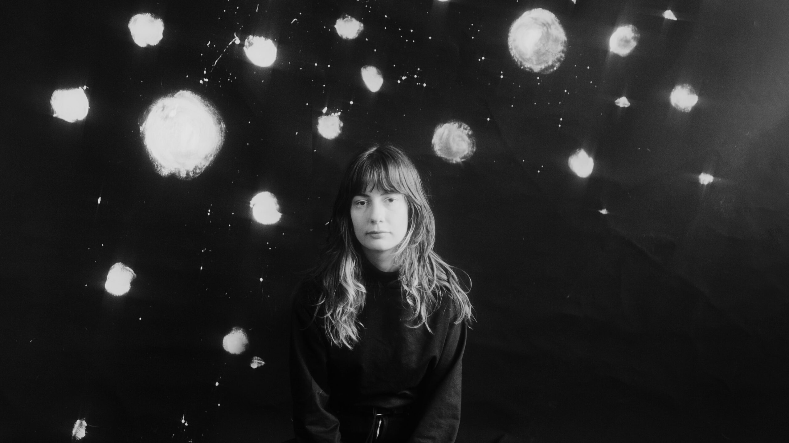 Sophie Lindinger sitzt vor einer Wand mit großen Farbklecksen. Das Bild ist Schwarz-Weiß.