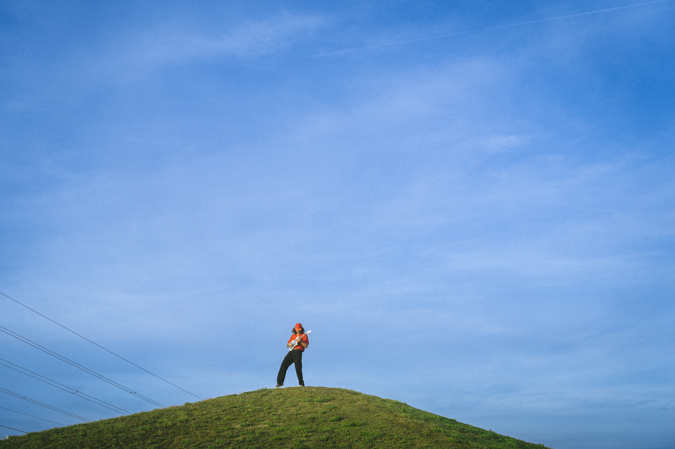 James Hersey steht mit einer Gitarre auf einem grünen Hügel. Der Himmel ist sehr hell und blau.