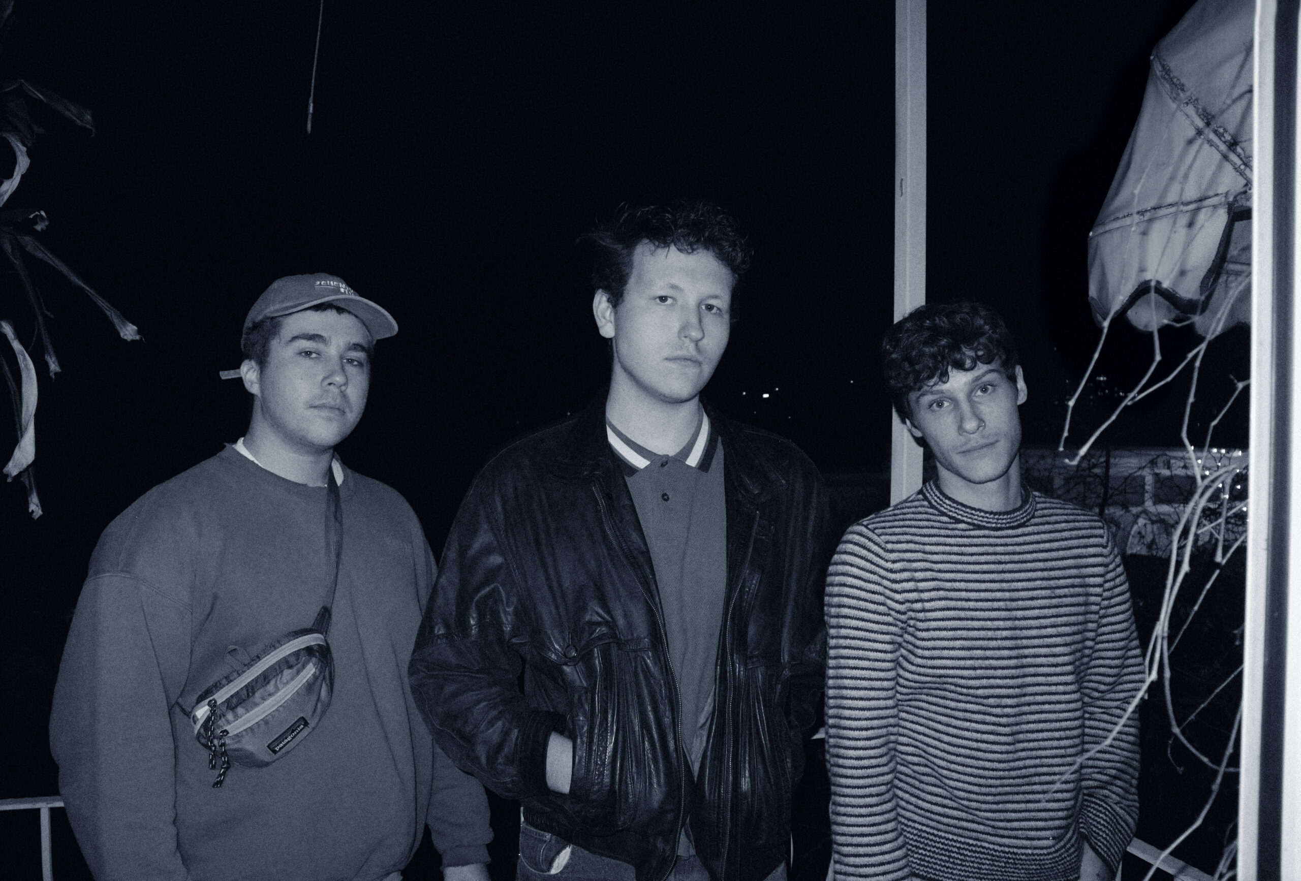 Auf dem Bild sind die drei Bandmember zu sehen: Links Basti, in der Mitte Jonas und rechts daneben steht Colin. ZU dritt stehen sie lässig auf einem Balkon, draußen ist es bereits dunkel.